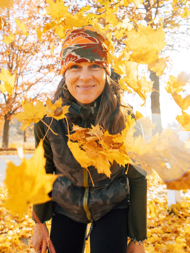 A woman smiling outdoors in autumn, surrounded by flying and fallen golden maple leaves. She is wearing a dark camouflage vest, dark pants, and a patterned beanie.